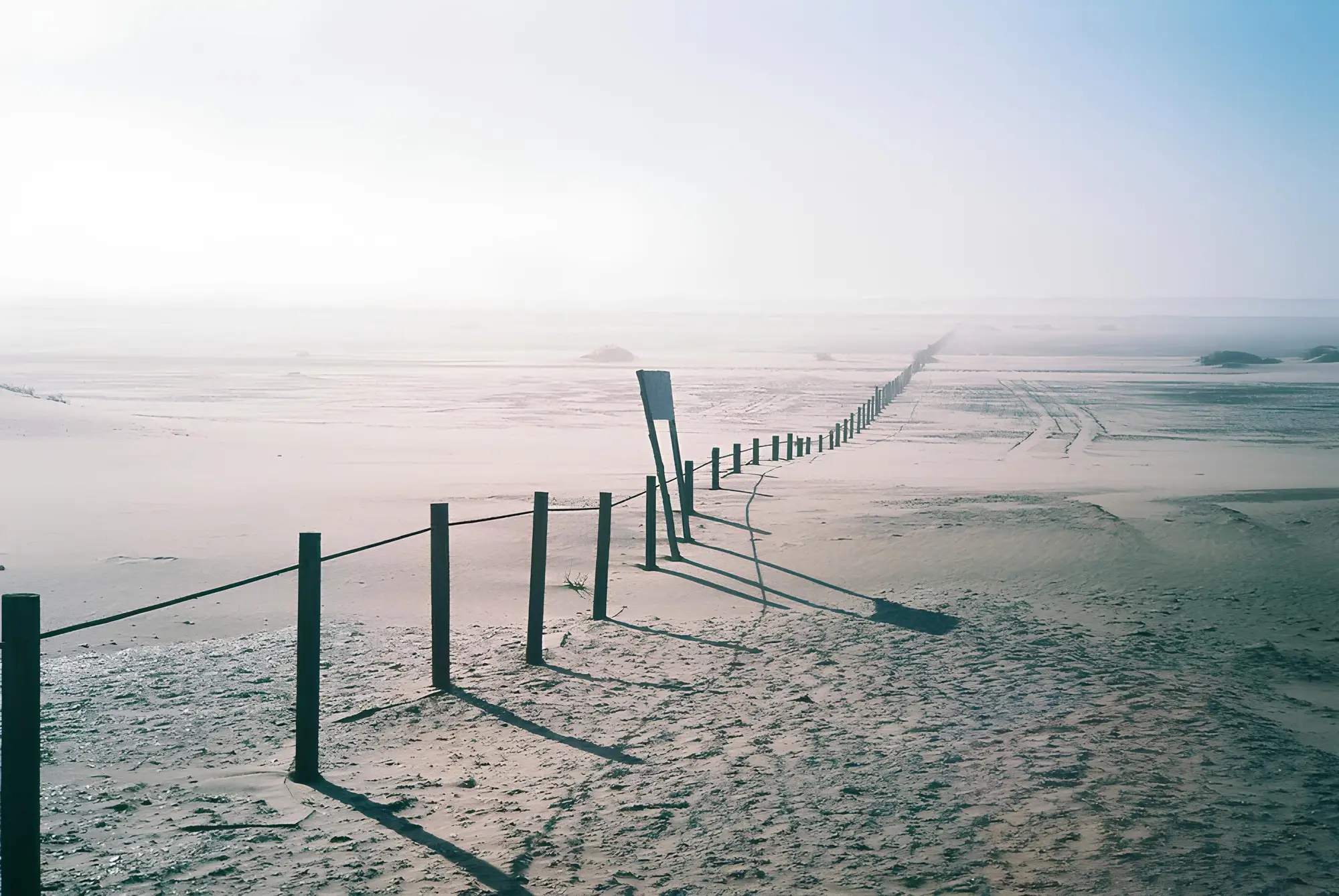A fence line of wooden posts running across open sand into haze